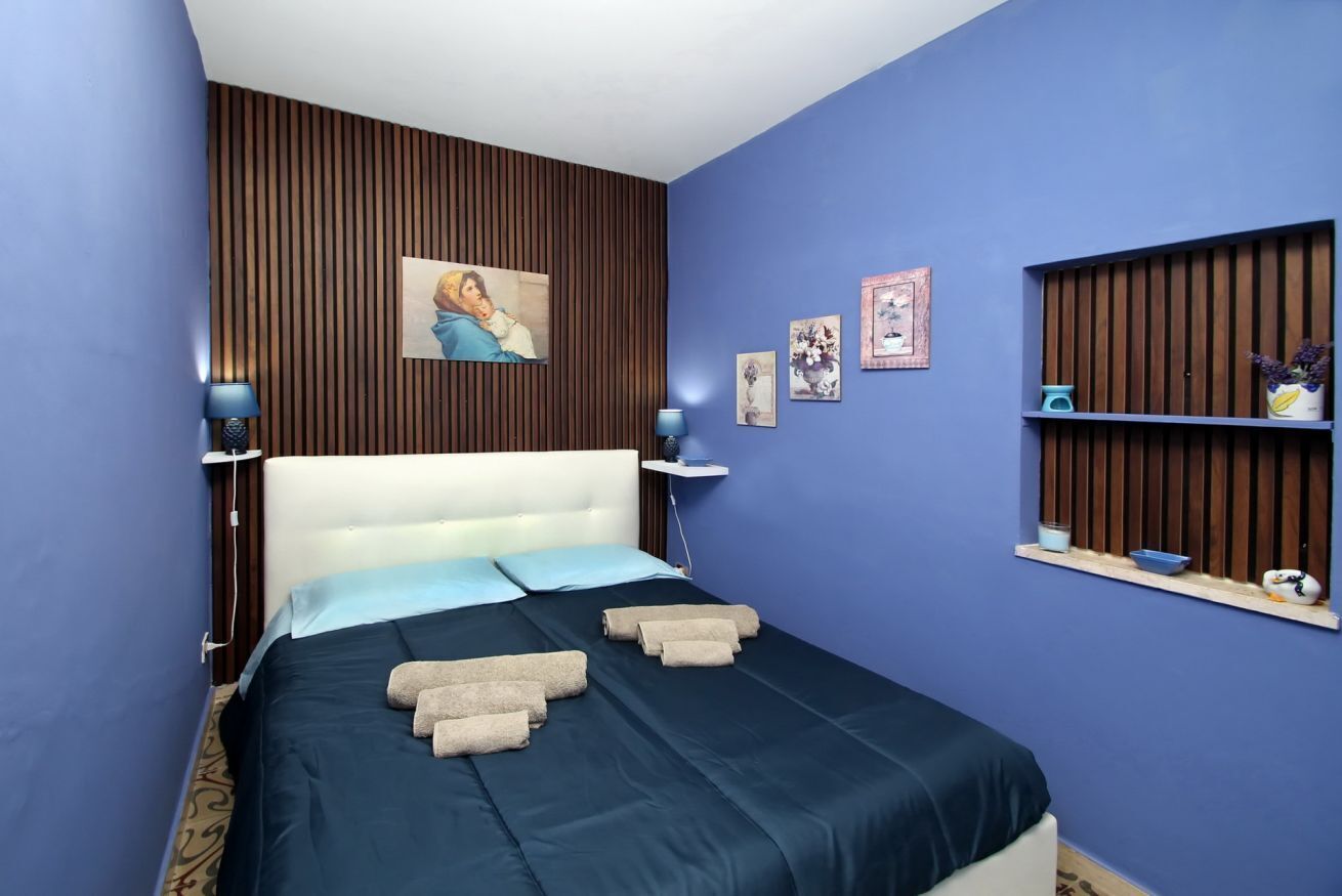 Un dormitorio ordenado con cama, lámparas de noche, estante en la pared con adornos y ventana con persianas.