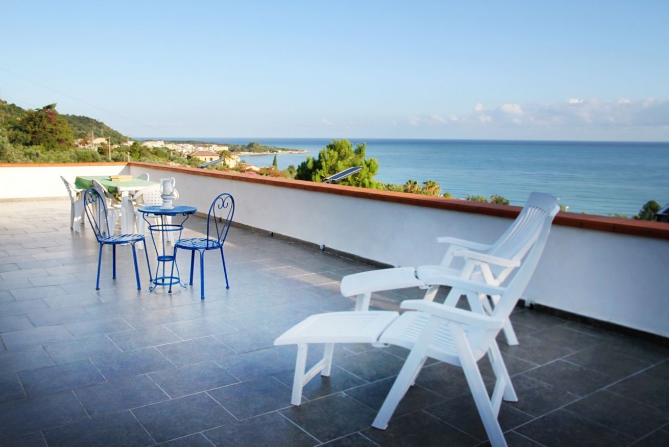 A terrace with white and blue chairs, a table, and a view of the sea.
