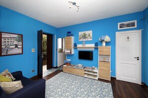 Ein Zimmer mit blauen Wänden, einem Holztischchen, einem an der Wand befestigten Fernseher und einer weißen Tür mit der Aufschrift 'Toilette'.