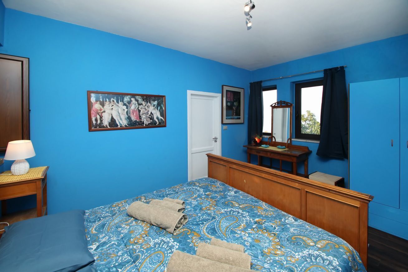 Ein Schlafzimmer mit blauer Wand, einem Bett mit gemustertem Überwurf, einem hölzernen Kopfteil, einem Nachttisch mit Lampe und einem Bild an der Wand.
