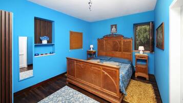 Une chambre avec un lit en bois, des murs bleus, une commode en bois, un miroir et une petite fenêtre avec une plante.