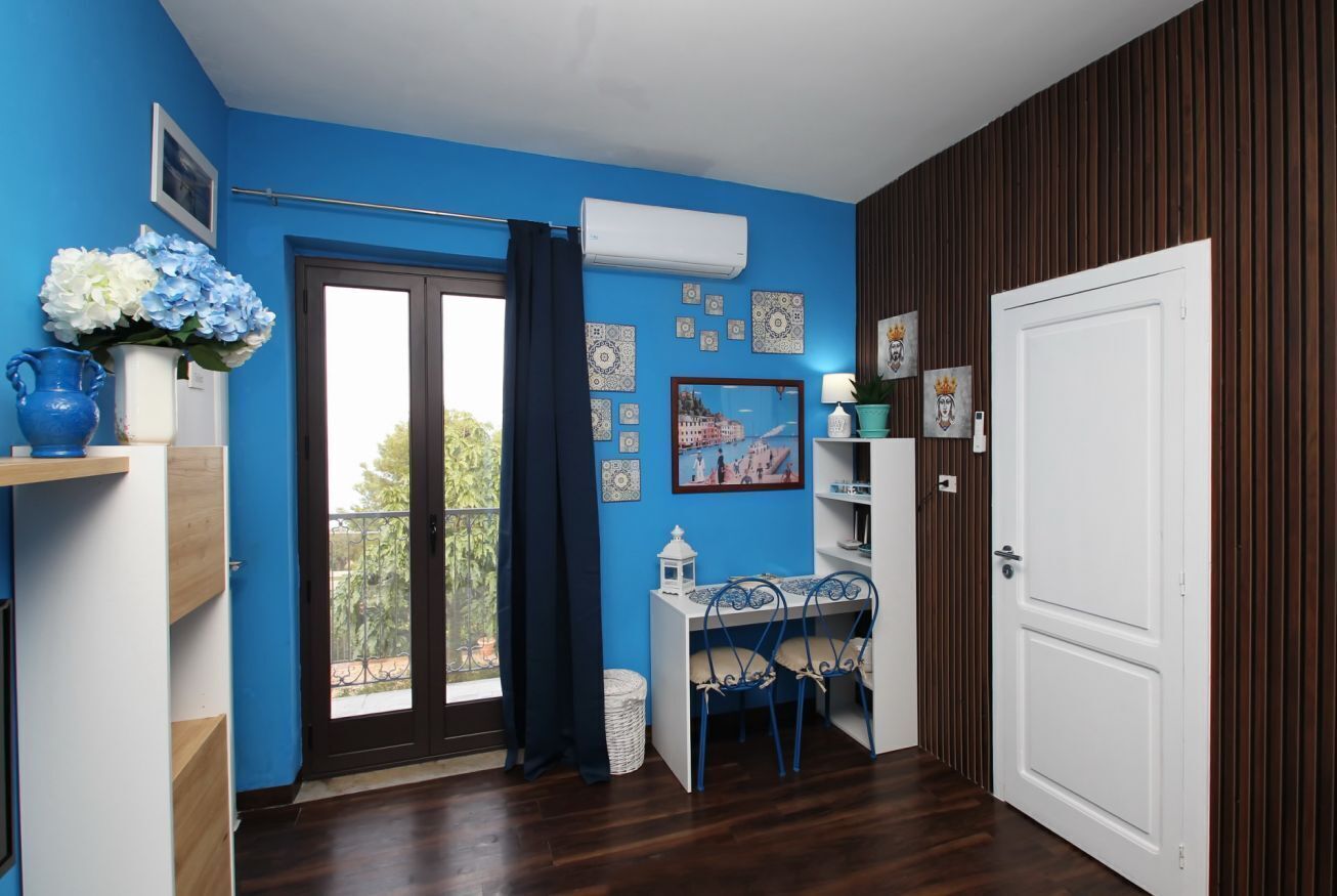 Um quarto com parede azul, piso de madeira, porta branca, janela com cortina, uma pequena mesa com cadeiras e um vaso com flores.