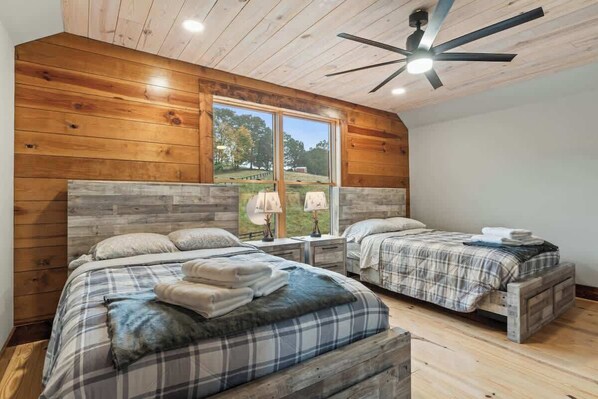 A bedroom with two beds, wooden walls, a large window, and a ceiling fan.
