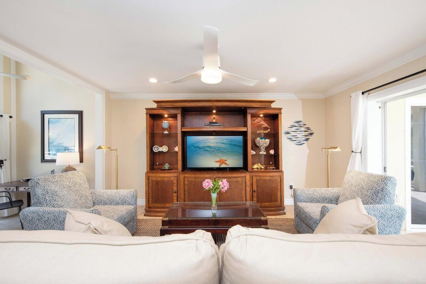 A living room with a large flat-screen TV, wooden entertainment unit, two patterned sofas, a coffee table with a vase, and a ceiling fan.