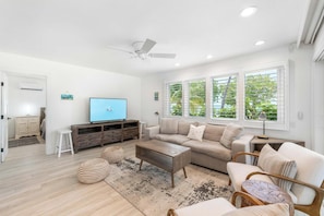 Ein modernes Wohnzimmer mit Sofa, Couchtisch und Fernseher, großen Fenstern mit Jalousien und einem Deckenventilator.