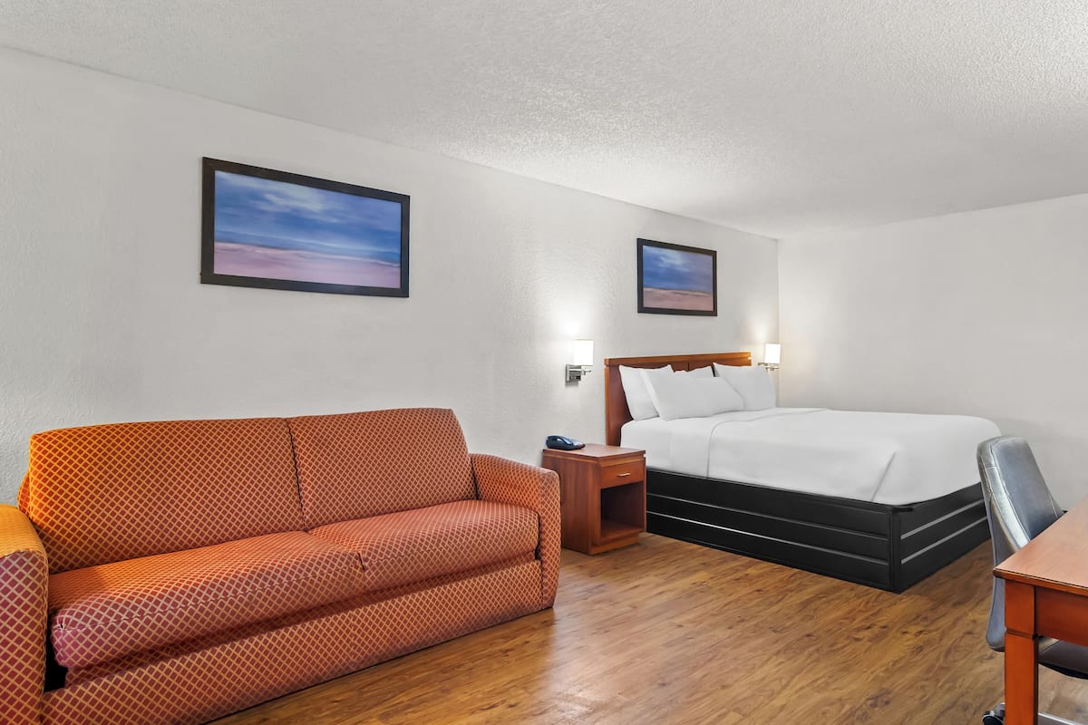 Habitación de hotel con suelo de madera, sofá, cama, mesita de noche y dos cuadros en la pared.