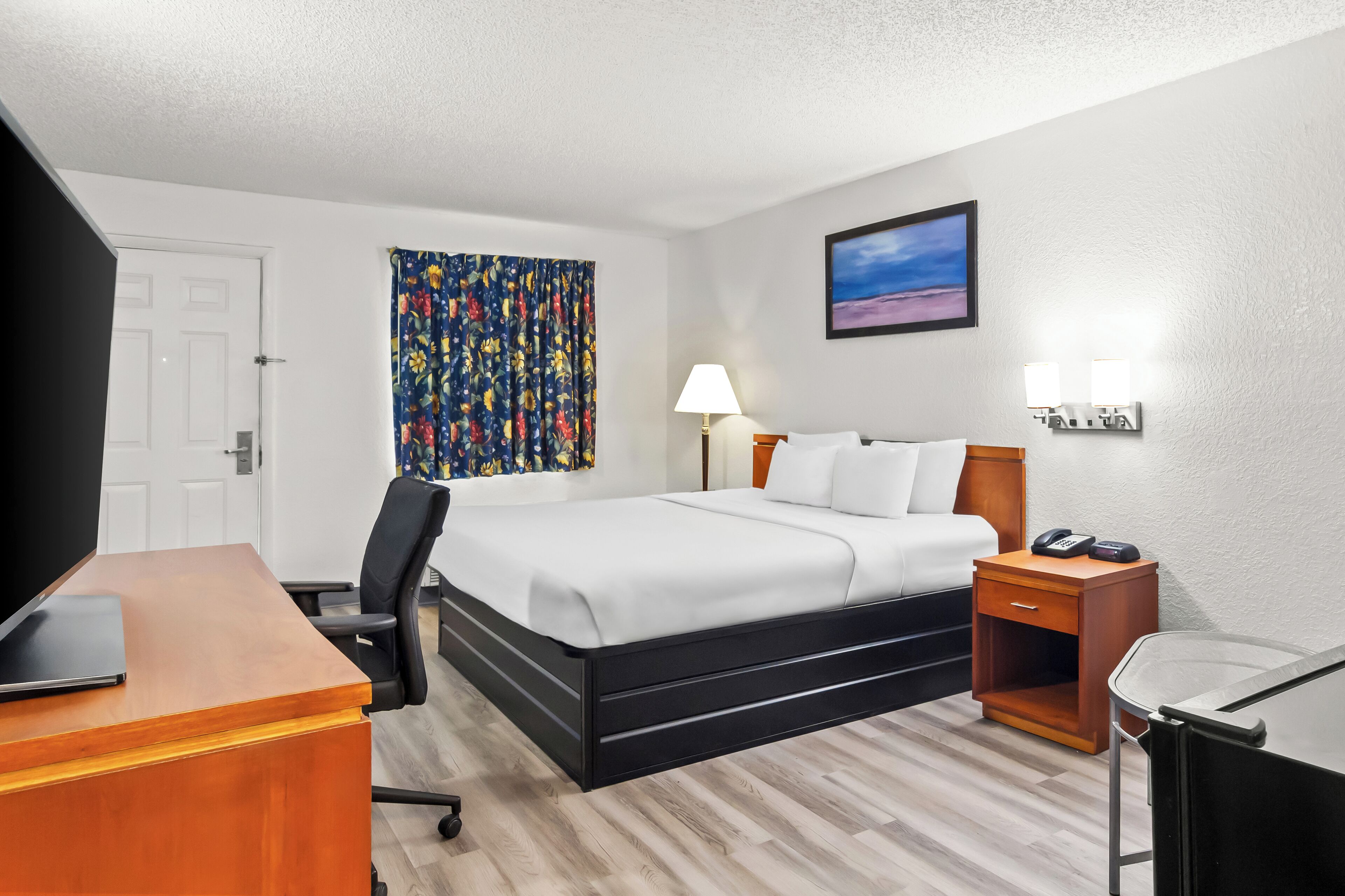 A hotel room with a bed, desk, chair, bedside table, lamp, and artwork on the wall.