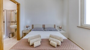 Un dormitorio con cama, mesitas de noche, una ventana y un baño visible a través de una puerta abierta.