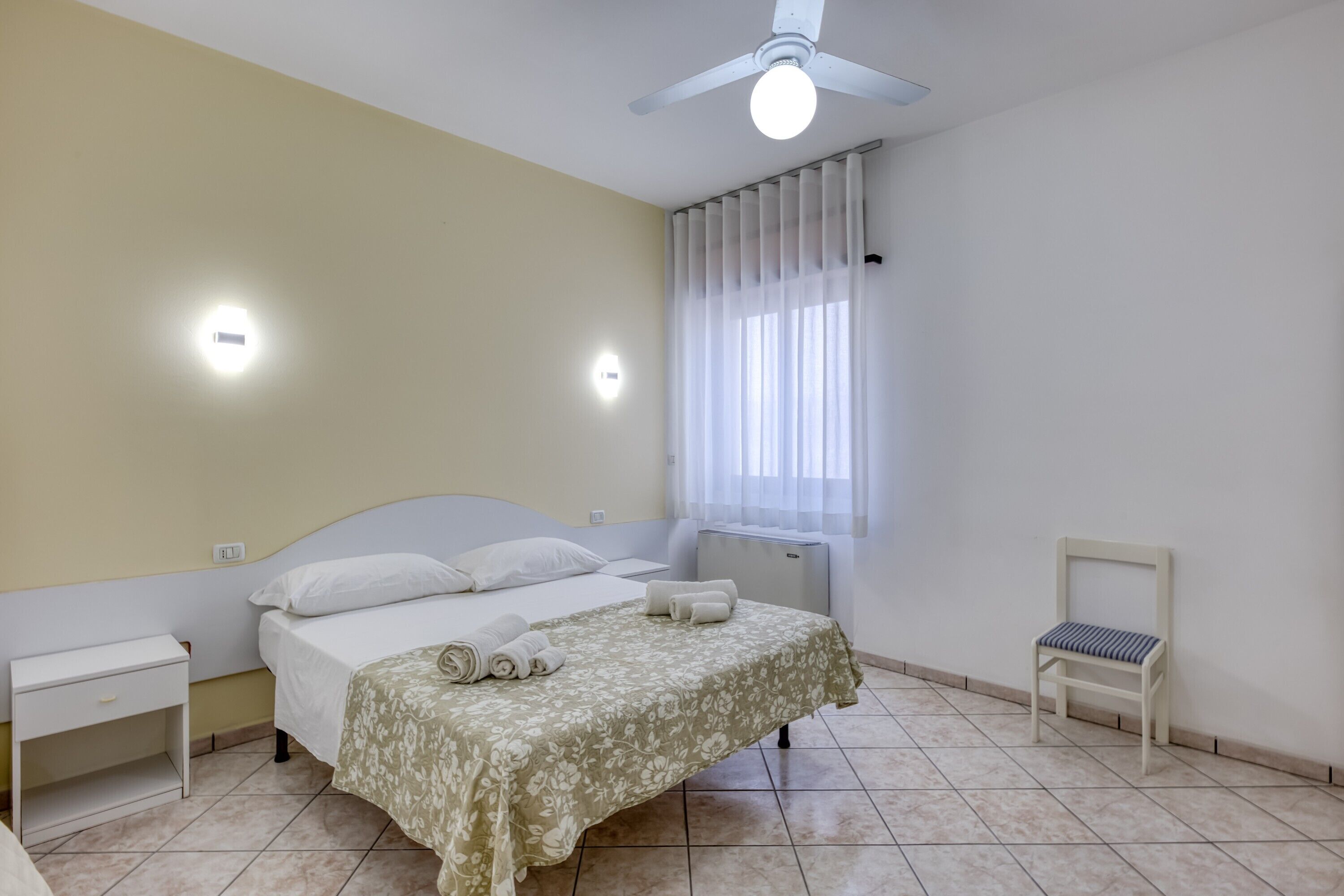 Een slaapkamer met een bed, nachtkastjes, een stoel, een plafondventilator en een raam met gordijnen.
