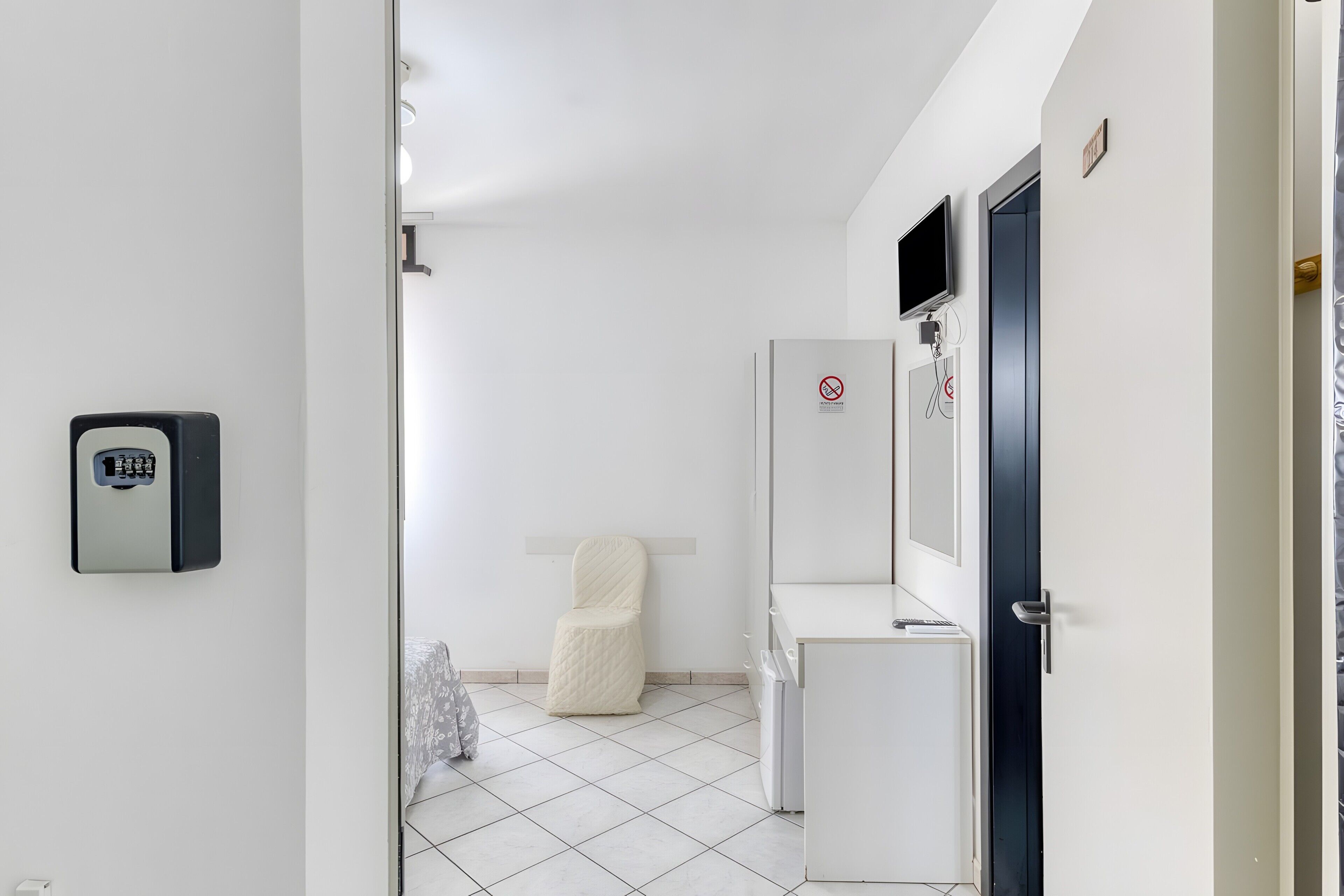 Une chambre d’hôtel avec un sol carrelé de carreaux blancs, un lit recouvert d’une couvre-lit à motifs, une armoire blanche, un petit meuble blanc et un téléviseur fixé au mur.