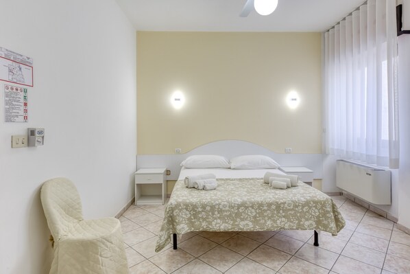 Un dormitorio con cama, una silla, dos mesitas de noche, aire acondicionado instalado en la pared y una ventana con cortinas.