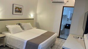 Et hotelværelse med en seng, et sengebord og et lille skrivebord med et spejl. Der er et billede på væggen, og en aircondition er monteret over sengen.