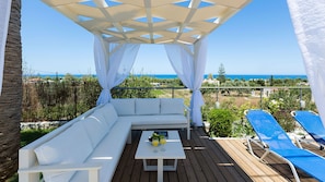 A covered outdoor seating area with a white sofa, a table with drinks, and blue lounge chairs. - Villa Aetos (Rethymno)