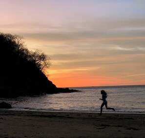 A beach scene with a person running along the shore during sunset.