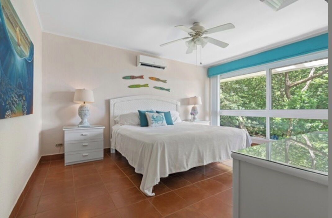 Une chambre à coucher avec un lit, des tables de chevet, un ventilateur de plafond et une fenêtre donnant sur des arbres.