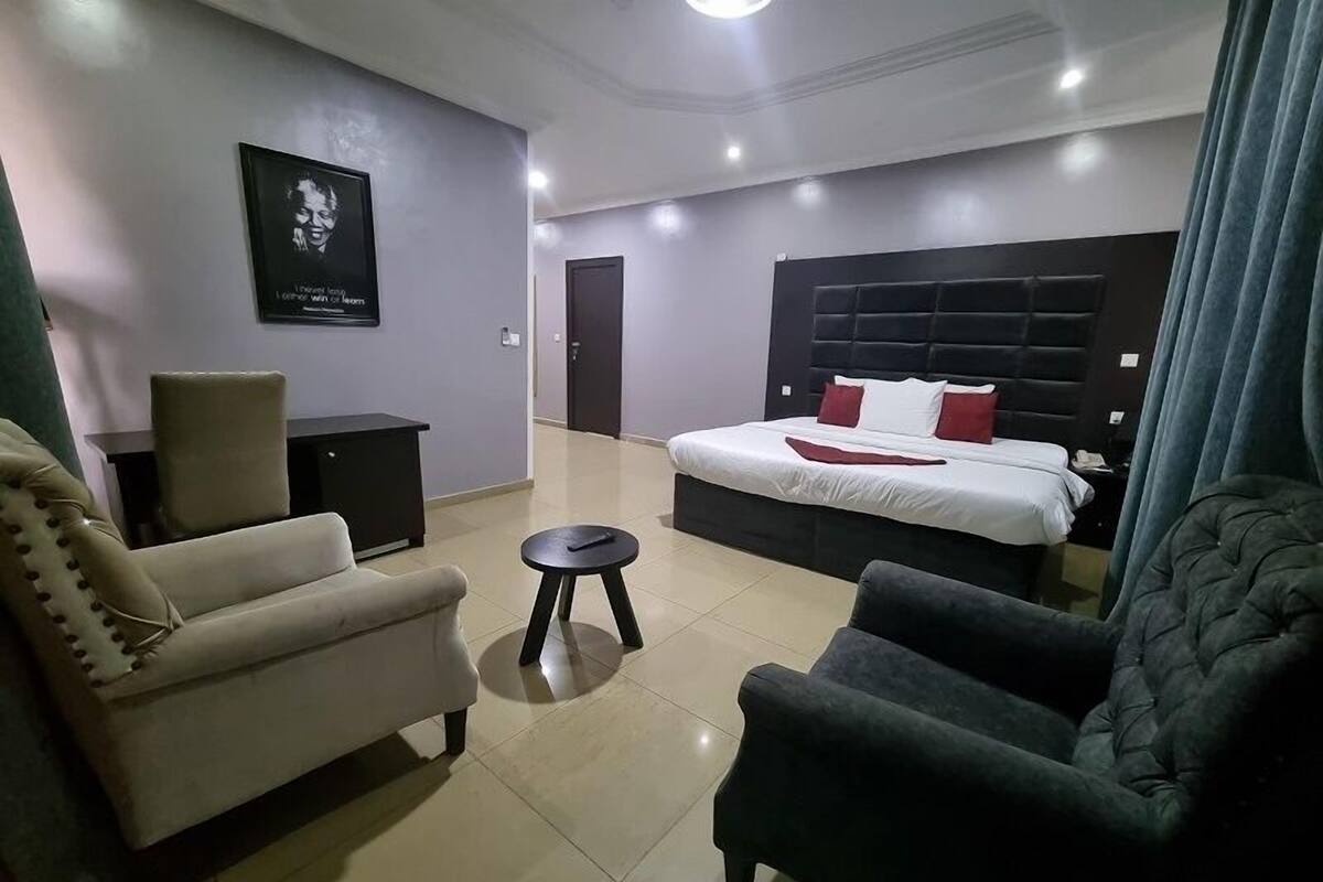 A modern hotel room with a large bed, a sofa, a small table, and a dark armchair.