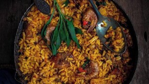 A large pot of rice with meat and vegetables, a spoon, and a rustic wooden table. - Hotel Guidotti (Abuja)