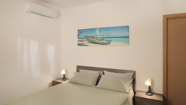 A bedroom with a bed, two bedside tables with lamps, a painting of a boat on the wall, and an air conditioning unit. - Villa Alice is an independent newly built villa, designed in the traditional “dammuso” style with its characteristic dome and overlooking the sea. It is a retreat immersed in nature, perfect for those seeking peace, privacy and an authentic stay. The (Marina di Caronia)