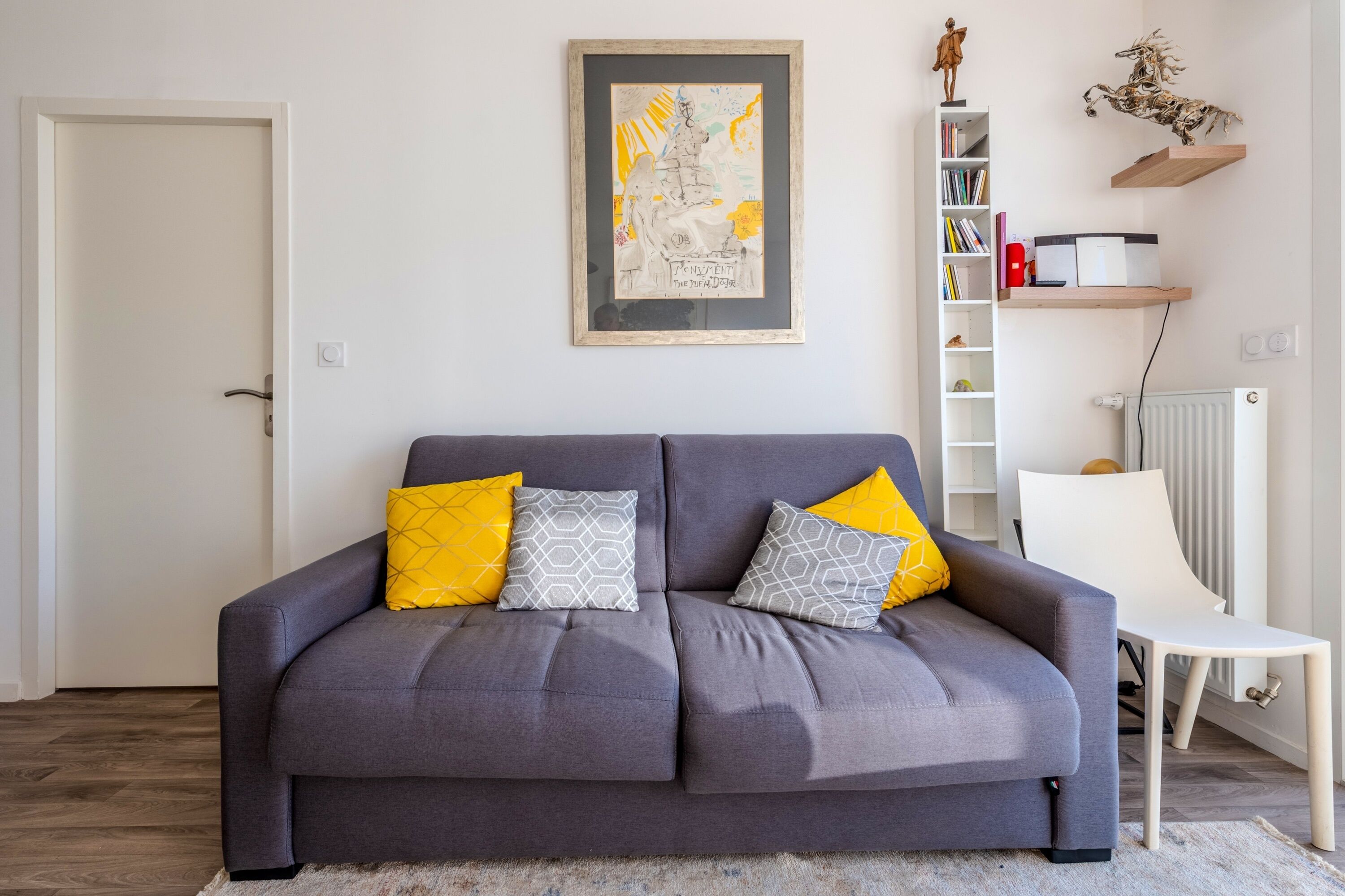 Un canapé gris agrémenté de deux coussins jaunes et de deux coussins gris à motifs, une chaise blanche, une bibliothèque remplie de livres et d’objets décoratifs, et une œuvre d’art encadrée accrochée au mur.