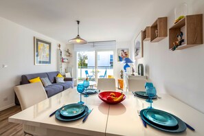 A modern living room with a sofa, dining table set with blue and red plates, and a kitchen area with wooden shelves. - Cap Lanroz - Avec terrasse (Brest)