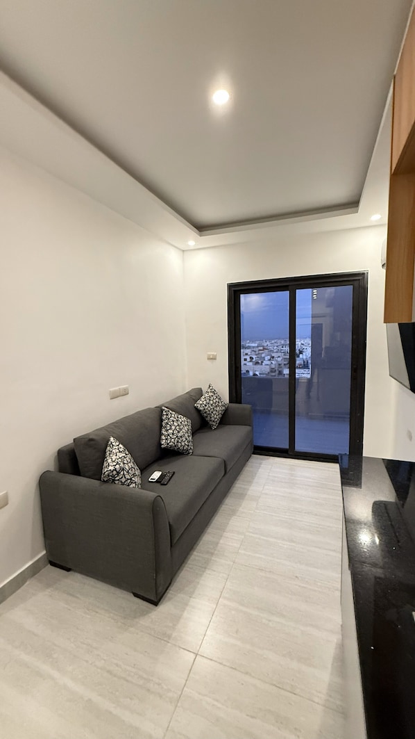 A modern living room with a grey sofa, patterned cushions, and a remote control on it. There is a large window with a view of a cityscape. - Riva suites (Irbid)