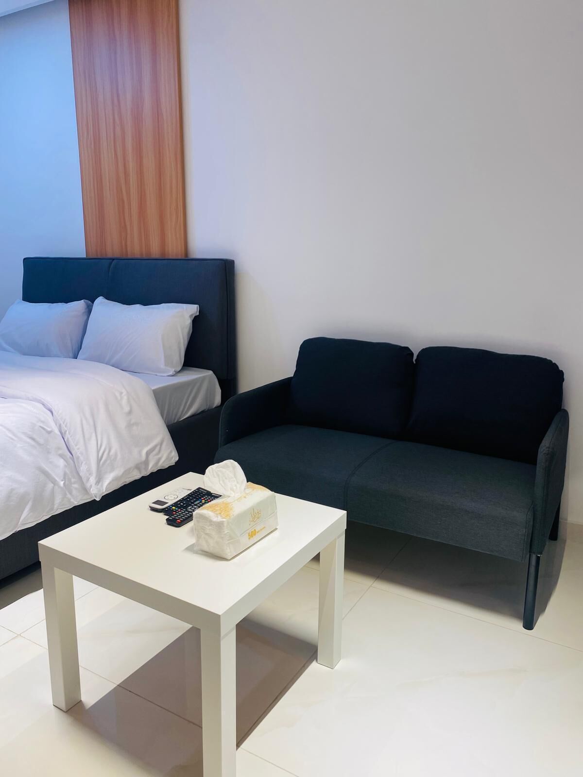 Ett modernt hotellrum med en säng, en mörk soffa och ett litet vitt bord med en fjärrkontroll och en ask med pappersnäsdukar.