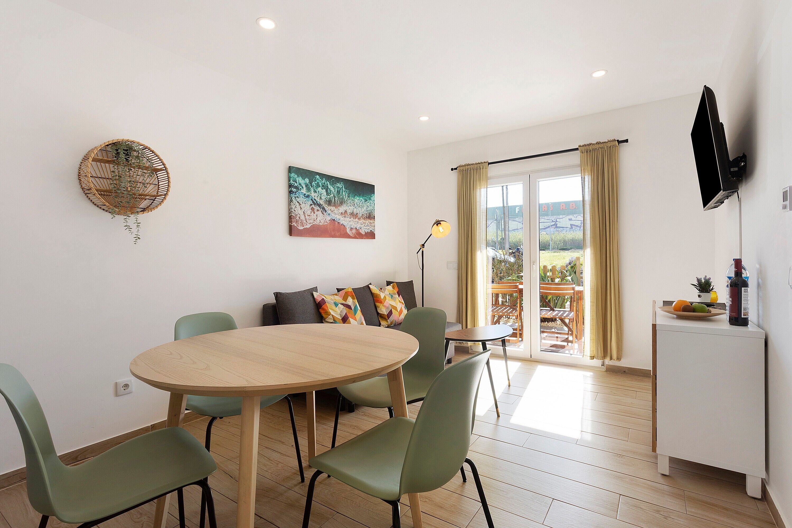 Un coin repas avec une table ronde en bois, des chaises vertes, un canapé agrémenté de coussins colorés, un tableau et un téléviseur fixé au mur.