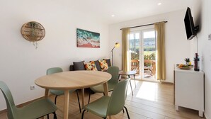 Un comedor con una mesa redonda de madera, sillas verdes, un sofá con cojines de colores, un cuadro y un televisor montado en la pared.