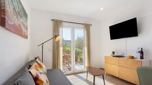 Una sala de estar con sofá, una mesa, un televisor montado en la pared, un ventanal corredizo que da a un área exterior y un armario de madera.