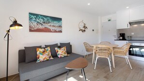 En moderne opholdsstue med sofa, lille bord, fletstole, et billede, en lampe og et tekøkken.