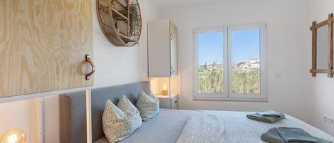 A bedroom with a bed, bedside table, lamp, wall decor, and a window with a view.