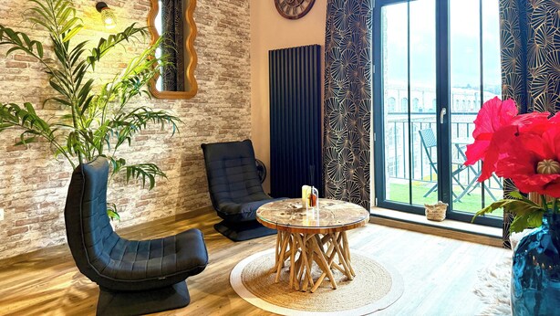 Een kamer met een bakstenen muur, een ronde houten tafel, twee zwarte stoelen, een groot raam met uitzicht en een kamerplant.