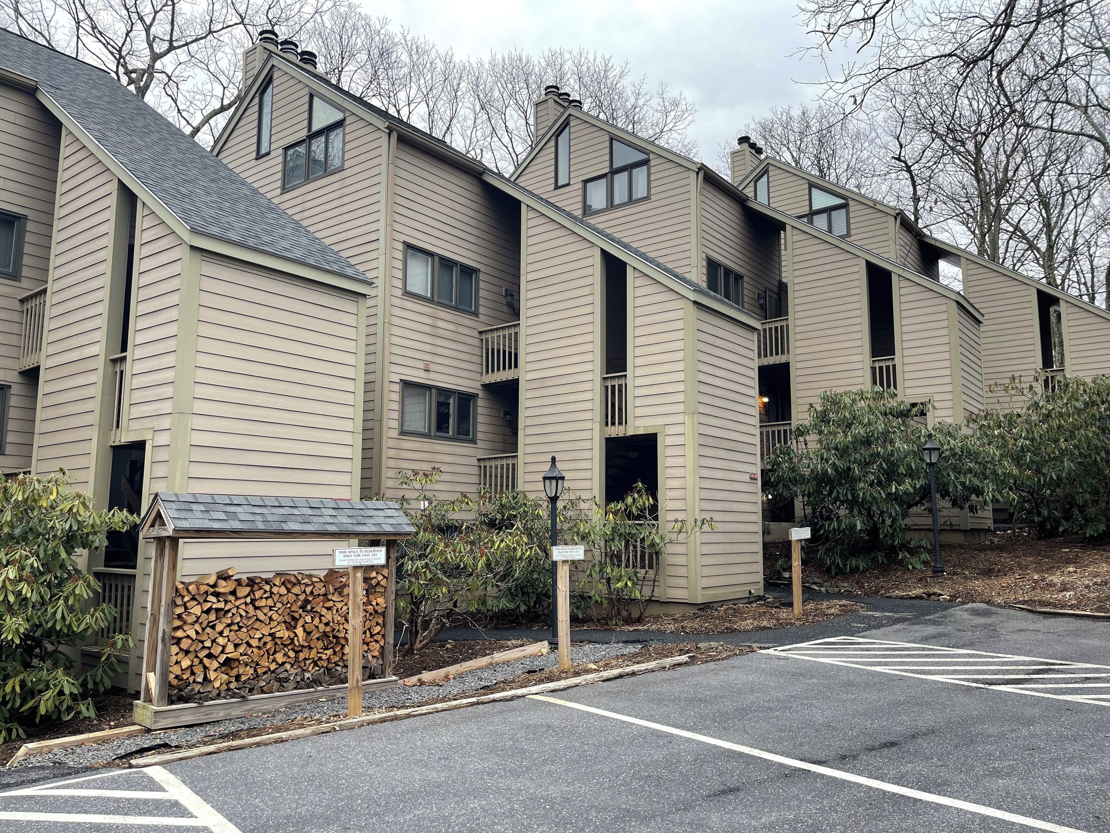 A row of beige multi-story buildings with balconies, a parking lot, and a stack of firewood.