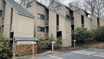 A row of beige multi-story buildings with balconies, a parking lot, and a stack of firewood.