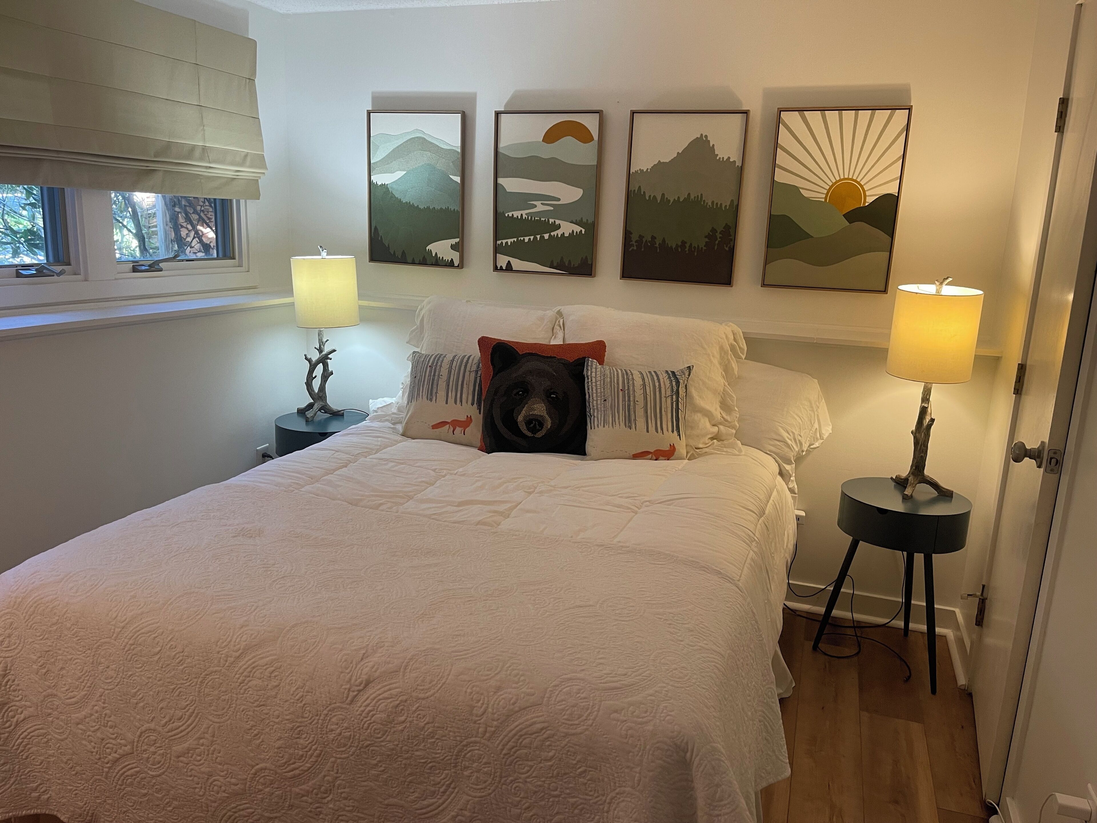 A bedroom with a bed, two bedside lamps, a window with blinds, and three framed landscape paintings on the wall.