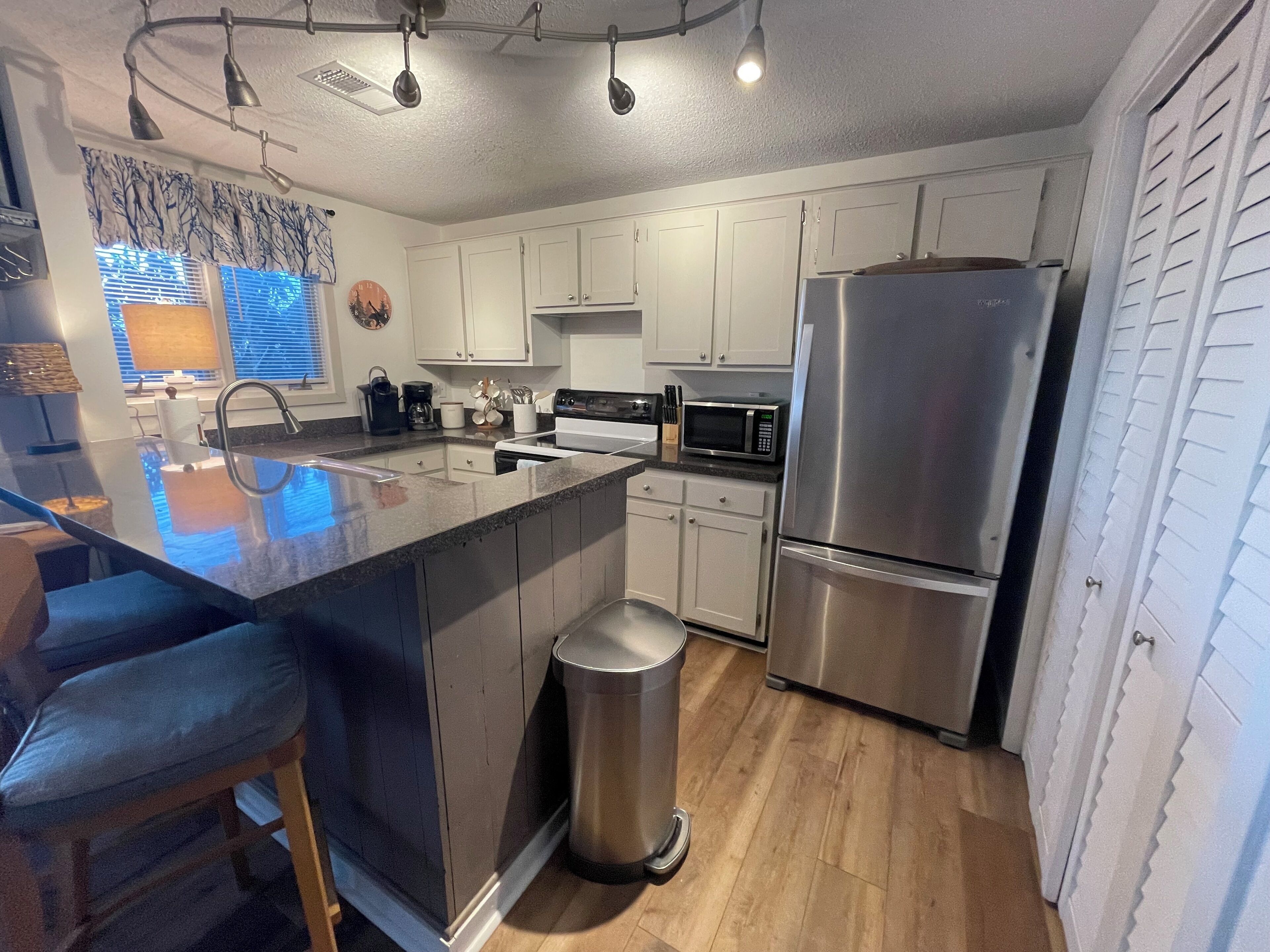 A modern kitchen with white cabinets, stainless steel appliances, a central island with bar stools, and a trash can.