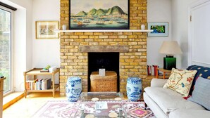 A living room with a brick fireplace, a painting above it, a bookshelf, and a sofa with decorative pillows. - Lovely 2 bed Flat With Garden in Queen's Park (London)