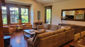 A living room with a large sofa, a coffee table, and a dining table. There are windows with a view of greenery outside. - Snowcreek Resort Vacation Rental 1513 (Mammoth Lakes)