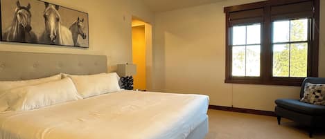 A bedroom with a large bed, a ceiling fan, a painting of horses, a window with blinds, and a chair with a pillow.
