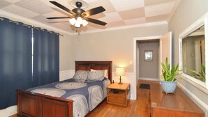 Un dormitorio con una cama de madera, un ventilador de techo, una mesita de noche con una lámpara y una planta en maceta.