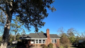 Una casa de ladrillo de un piso con chimenea, rodeada de un patio cubierto de césped y árboles.