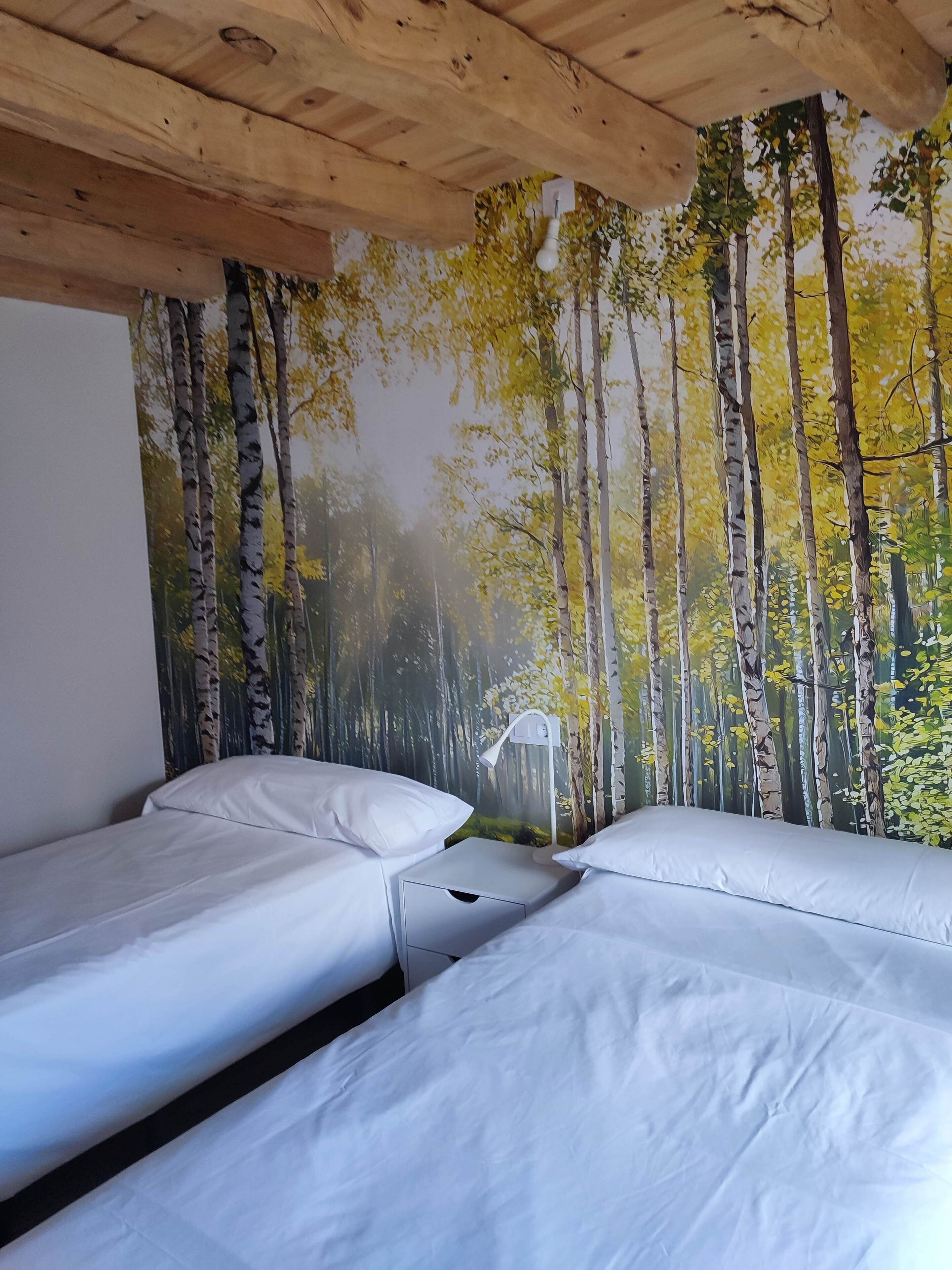 A room with two beds, a wooden ceiling, and a forest-themed wall mural.