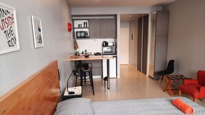 Una camera d'albergo compatta con un letto, una scrivania con una sedia, angolo cottura e una poltrona rossa.