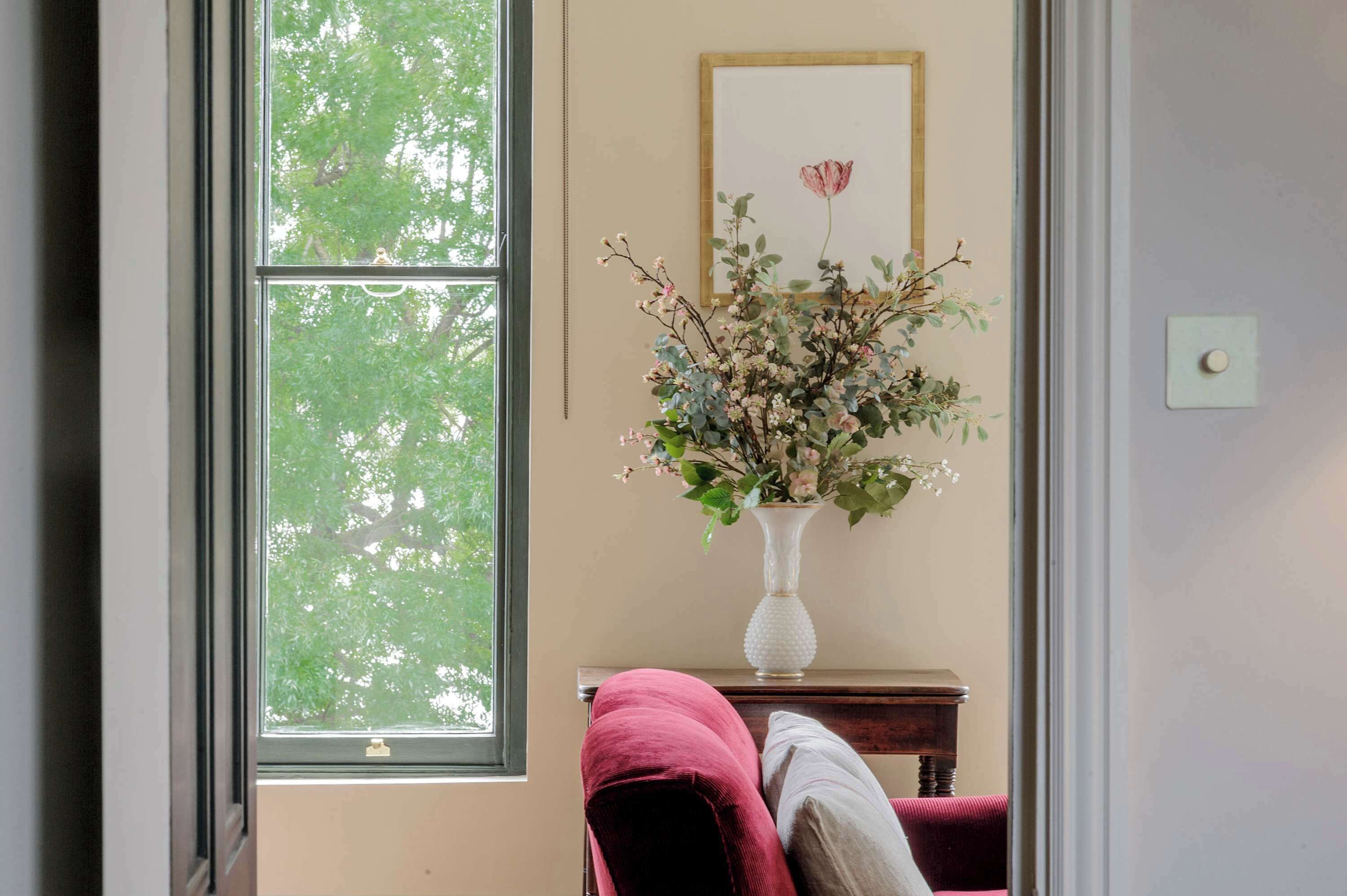A room with a large window, a vase with flowers, a framed picture, and a small table with a cushion.