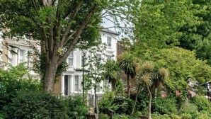 A well-maintained garden with a bench, a large tree, and a white building with shutters in the background. - Stylish 1 Bedroom Apartment - Notting Hill (London)