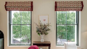 A room with two large windows, a vase with flowers on a small table, a red sofa, and a white armchair. - Stylish 1 Bedroom Apartment - Notting Hill (London)