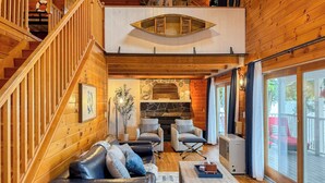 Een ruime woonkamer met een houten trap, een stenen open haard en een groot raam met uitzicht op de omgeving.