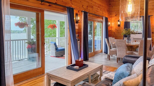 Een gezellig cabin interieur met houten vloer, een leren bank, een eettafel en een balkon met uitzicht op een meer en ligstoelen.