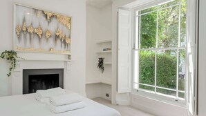 Une chambre à coucher avec un lit, une cheminée, une grande fenêtre et une étagère fixée au mur, où sont rangés des livres.