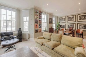 Een ruime woonkamer met een groot raam, een zwarte bureaustoel, een beige bank en een eethoek met tafel en stoelen.